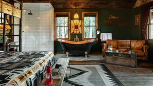 A rustic cabin interior with a large bed, a clawfoot bathtub, a leather sofa, and a wooden coffee table.