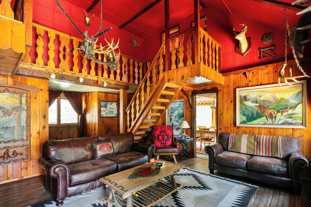 A rustic interior with wooden walls, a staircase, leather sofas, and a painting of deer.