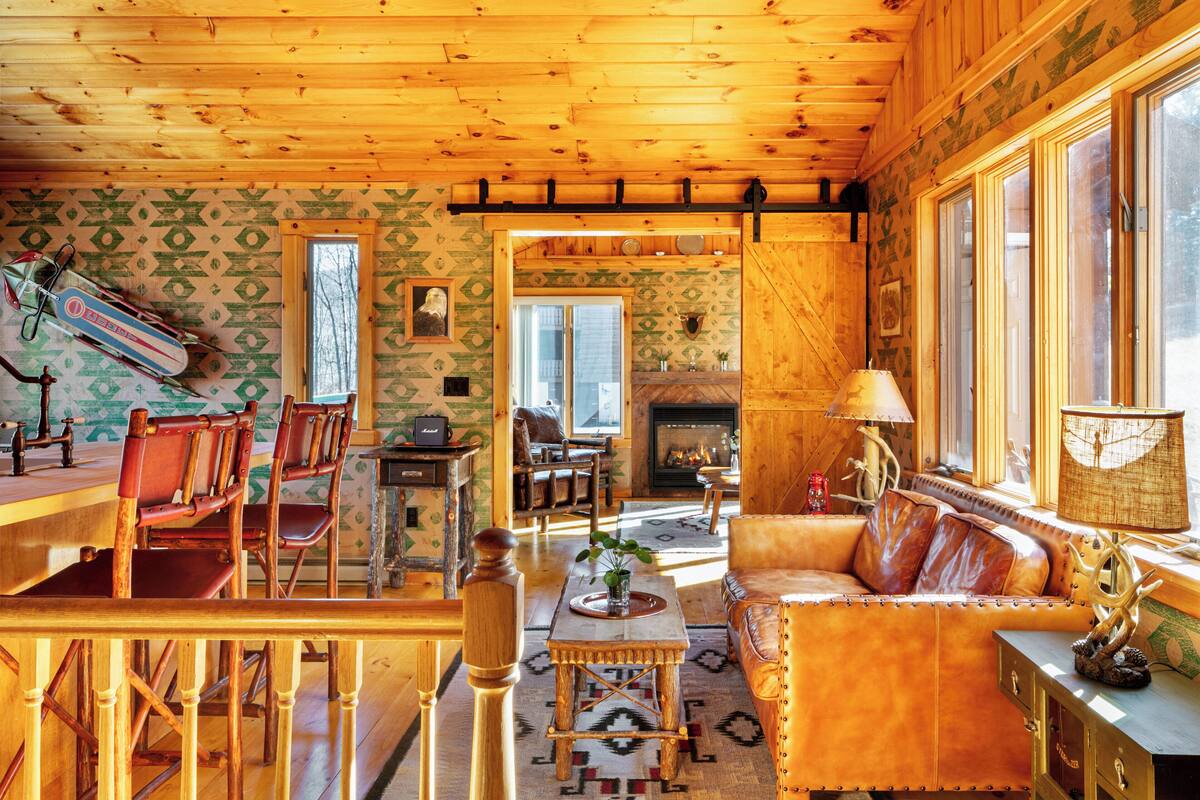 A cozy cabin interior with a wooden dining area, a fireplace, and a seating area with a sofa and chairs.