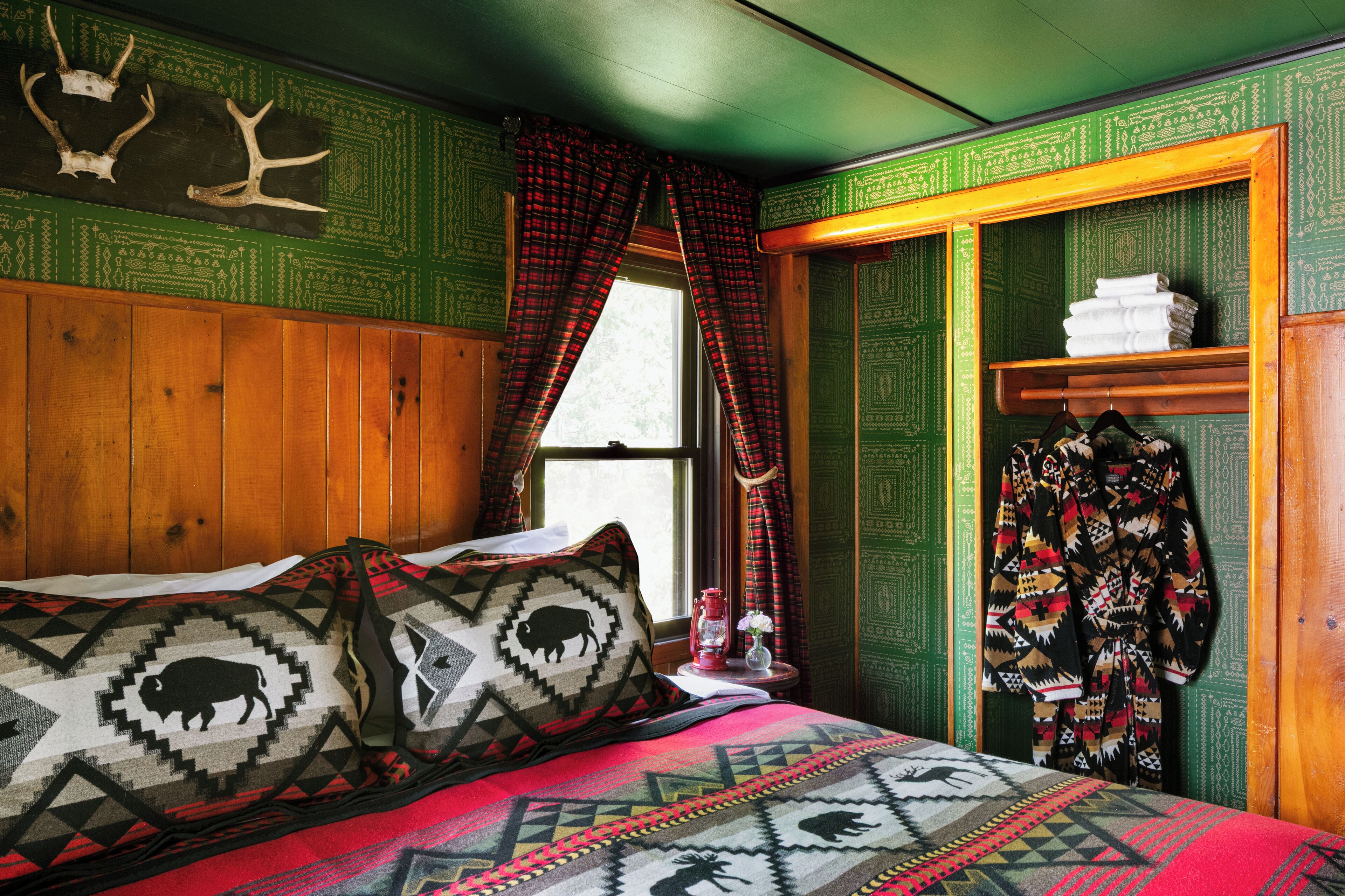 Una habitación con una cama, almohadas estampadas, una ventana con cortinas, un armario de madera y un estante con toallas dobladas.