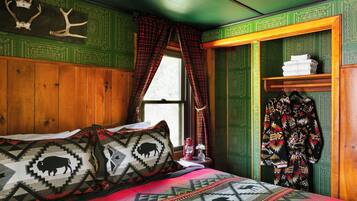 Una habitación con una cama, almohadas estampadas, una ventana con cortinas, un armario de madera y un estante con toallas dobladas.