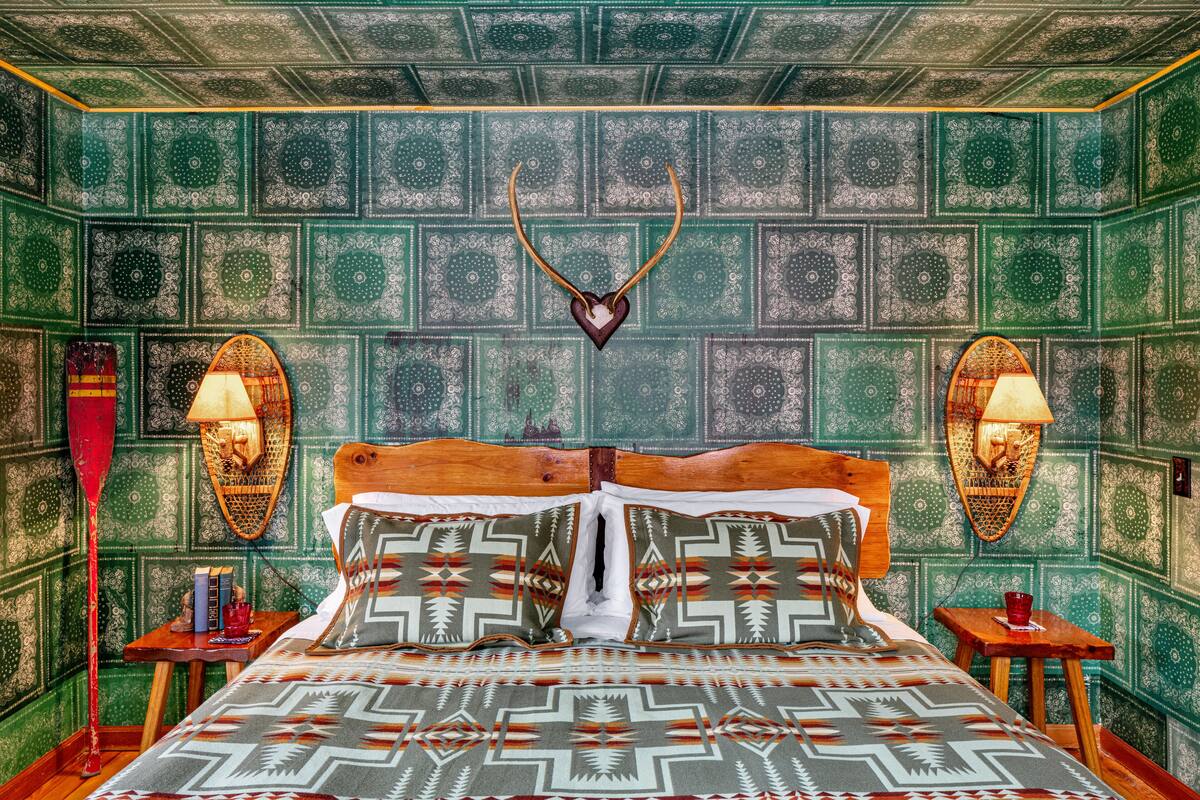 A room with a patterned green wall, a wooden bed with patterned pillows, a mounted antler, and two wall-mounted lamps.