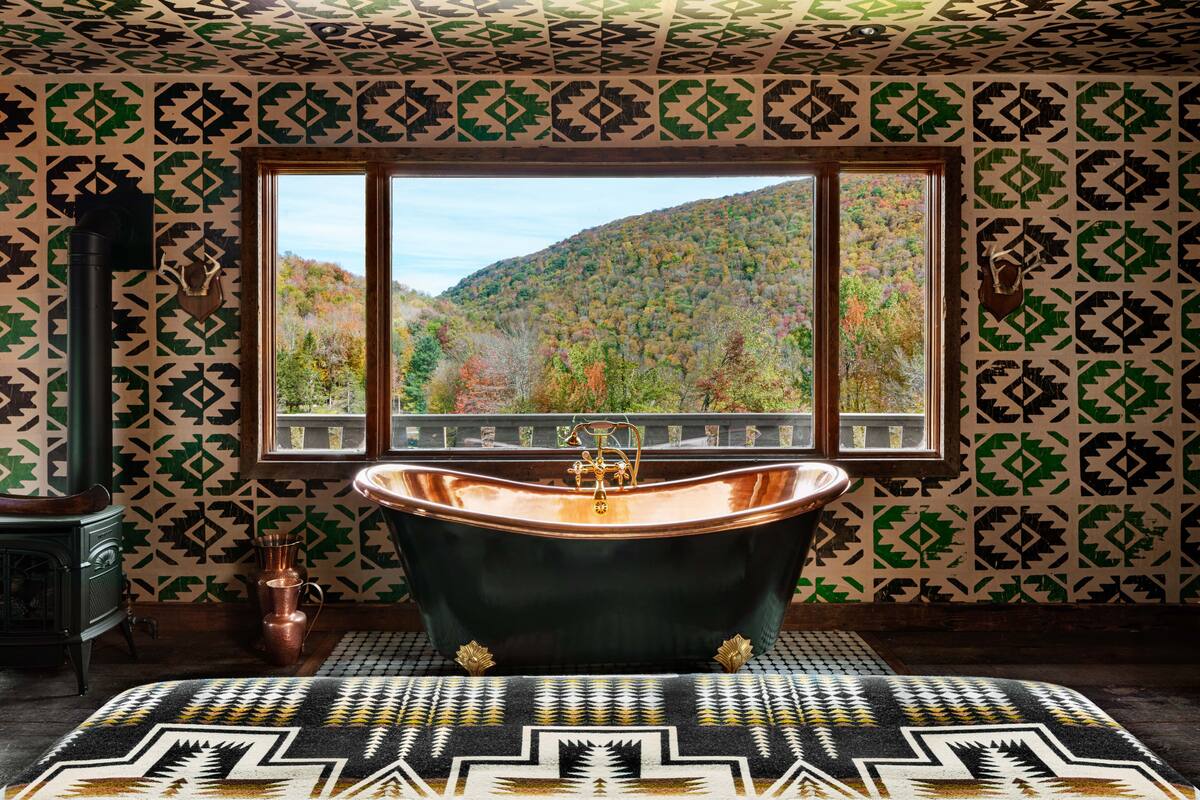 A bathroom with a freestanding bathtub, a window with a view of a forested hillside, patterned wallpaper, and a patterned rug.