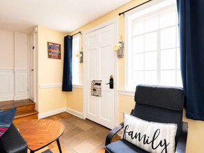 A room with a wooden floor, a black chair with a 'family' pillow, a small round table, a door with a black handle, and a welcome sign on the wall.
