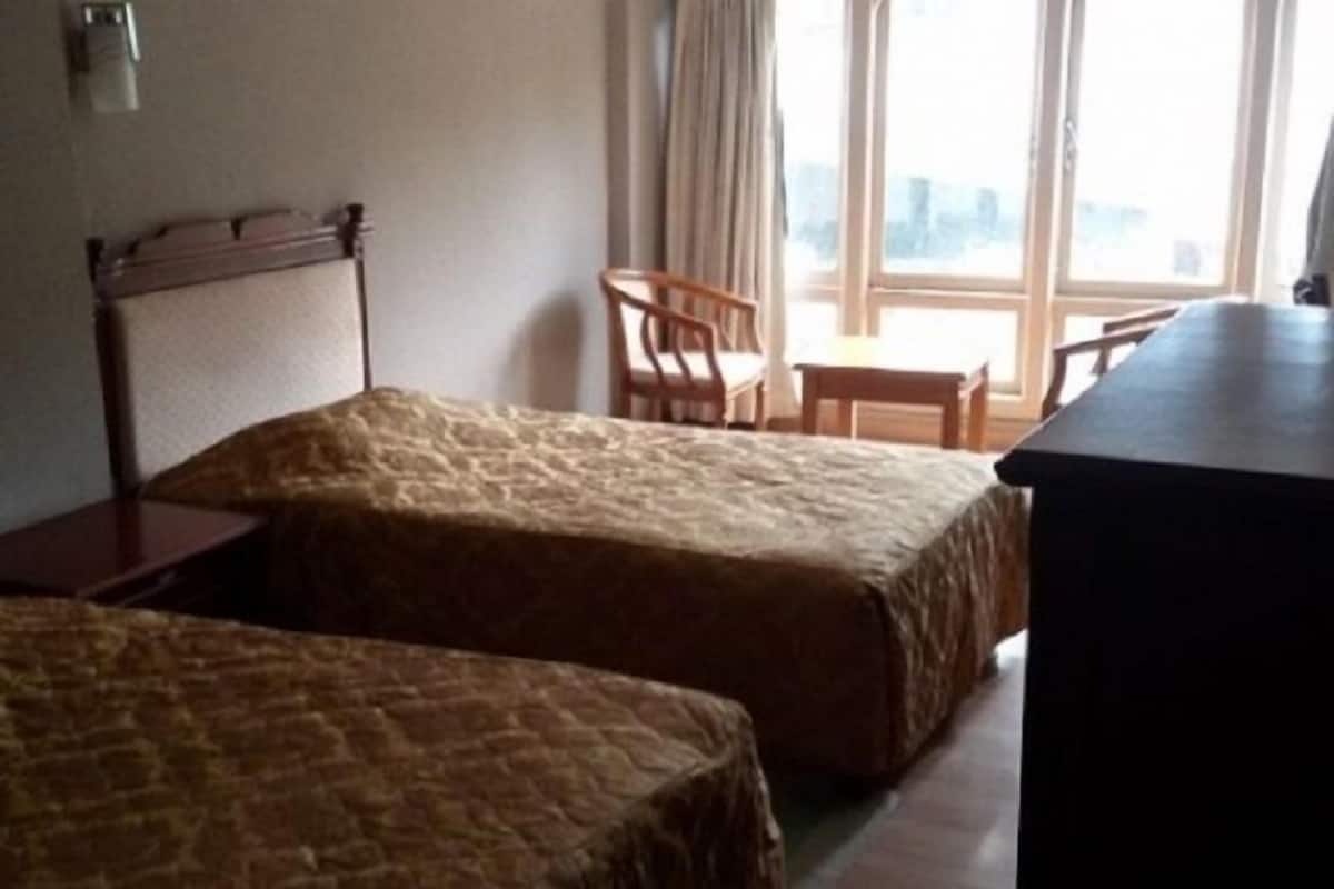 A hotel room with two single beds, a wooden headboard, a small table, a chair, and a large window with a view.