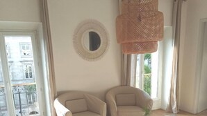 Un salon spacieux avec deux fauteuils beiges, une table basse en verre et un luminaire suspendu en fibres tressées. Un grand miroir rond est accroché au mur, et une grande fenêtre offre une vue sur l’extérieur.