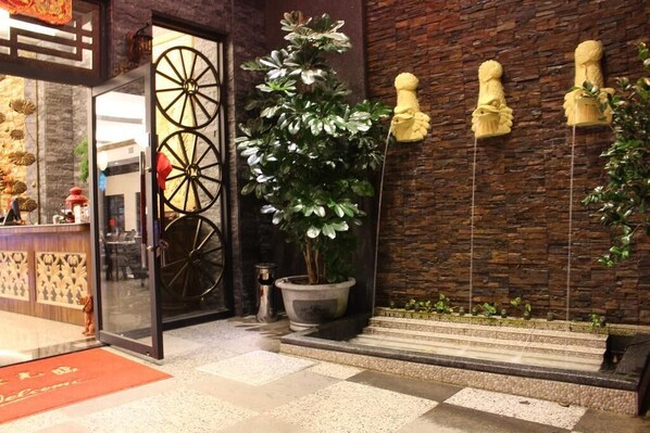 Entrada de hotel con una planta en maceta grande, puerta de cristal y una pared con esculturas decorativas.