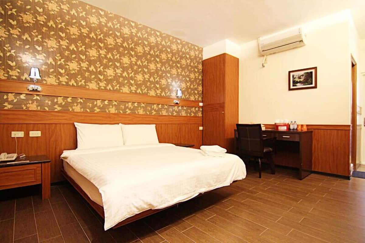 A hotel room with a large bed, a desk, and a chair. There is a wooden headboard, a wall-mounted lamp, and an air conditioning unit.
