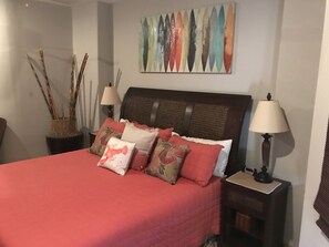 Un lit bien fait, avec une couvre-lit rouge, plusieurs oreillers et une tête de lit. On y trouve deux tables de chevet avec des lampes, une œuvre d’art murale au-dessus du lit, et un vase décoratif avec des branches sur le côté gauche.