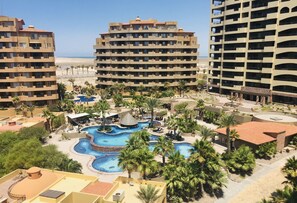 Resort com vários edifícios, piscina central e palmeiras.