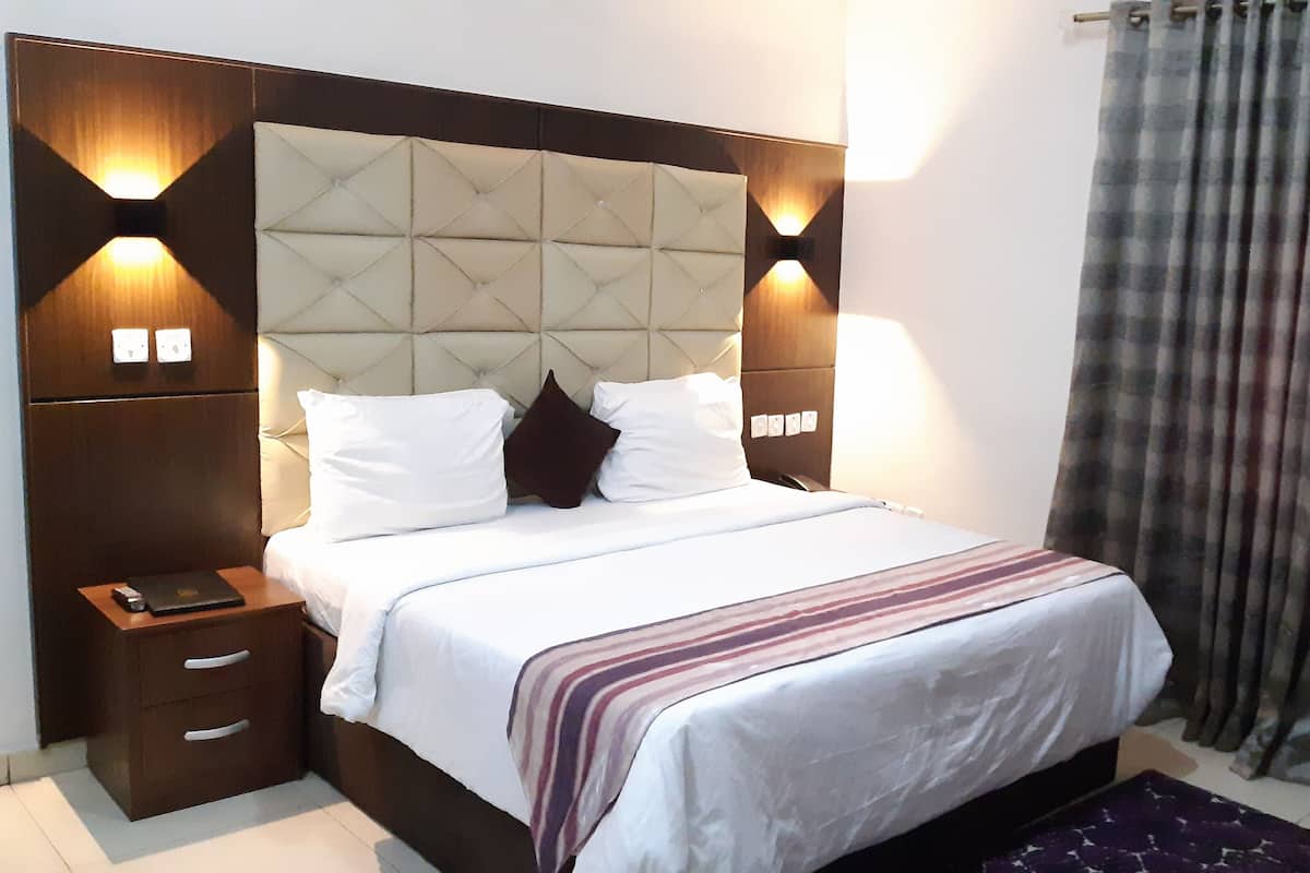 A neatly made bed with a tufted headboard, white bedding, and a striped throw. There is a nightstand with a phone and a remote control. The room has a dark wooden headboard and wall sconces.