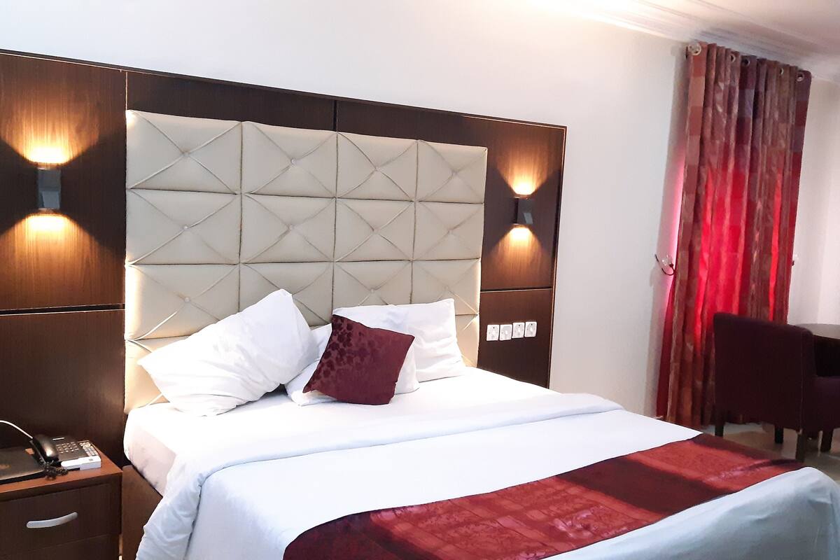 A hotel room with a large bed, a wooden nightstand with a telephone and a laptop, a red and white patterned bedspread, and a red curtain.