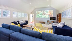 A spacious living room with a large screen displaying a nature scene, a dark blue sofa set, a wooden coffee table, and a bookshelf.