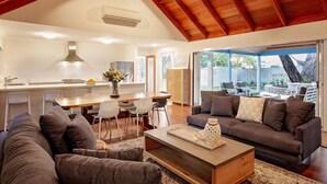 A spacious living area with a high ceiling, a dining area with a wooden table and chairs, and a view of the backyard through large sliding glass doors.