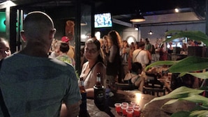 Um bar movimentado com pessoas em pé e sentadas, uma televisão ao fundo e um balcão cheio de bebidas.