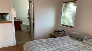 Un dormitorio con cama, mesita de noche, ventana con persianas y puerta que da a un baño.