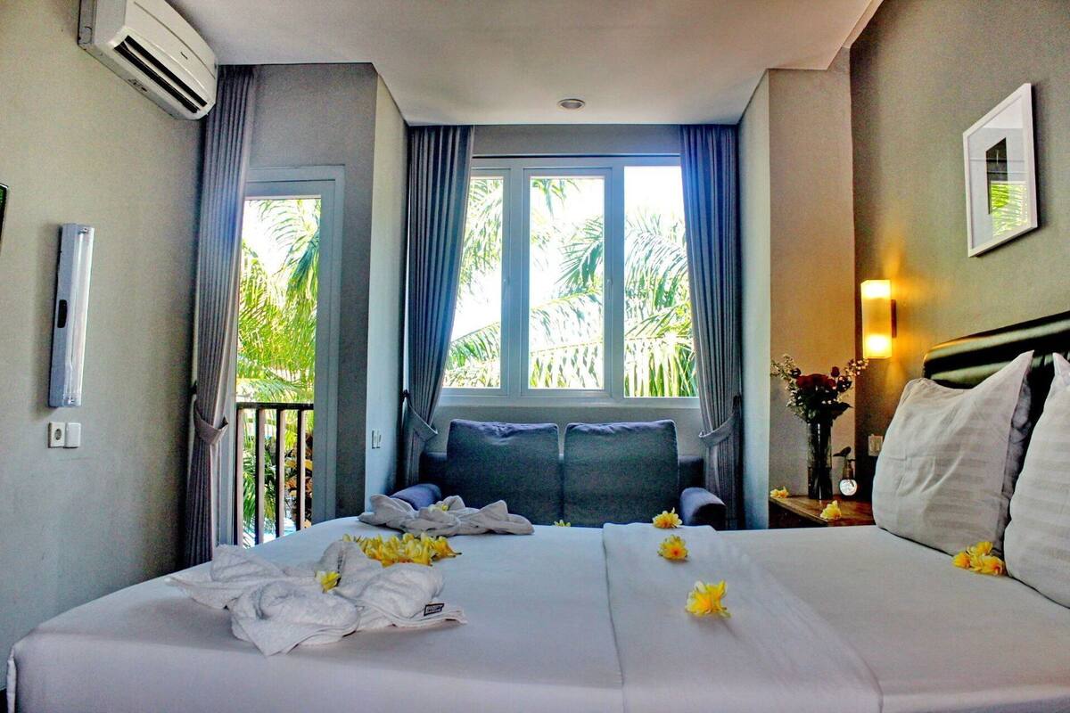 Habitación de hotel con cama, cortinas, ventana con vistas a palmeras, unidad de aire acondicionado y mesita de noche con un jarrón de flores.