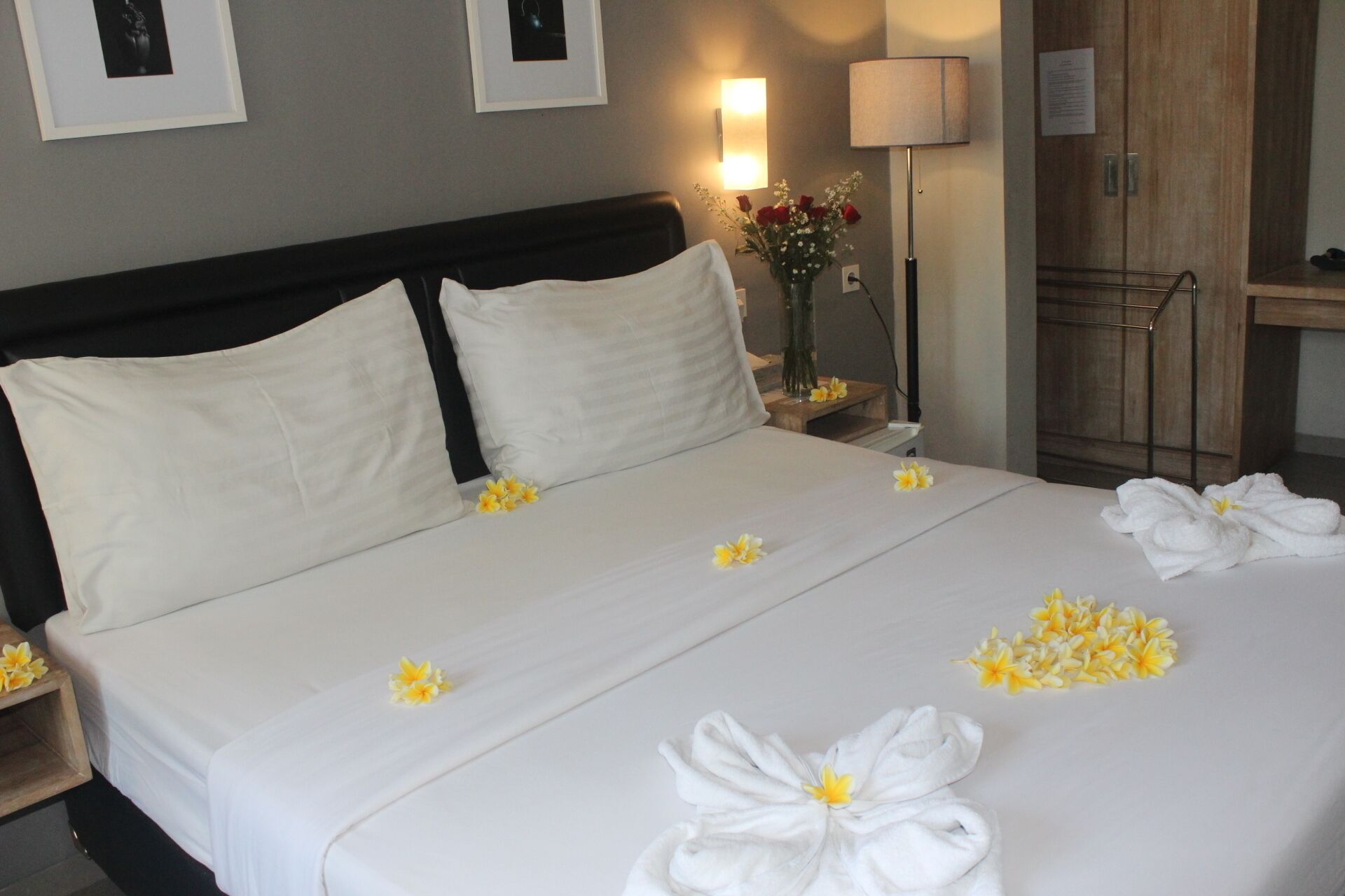 A hotel room with a large bed, two pillows, a nightstand with a lamp, a vase with flowers, and a wooden cabinet.