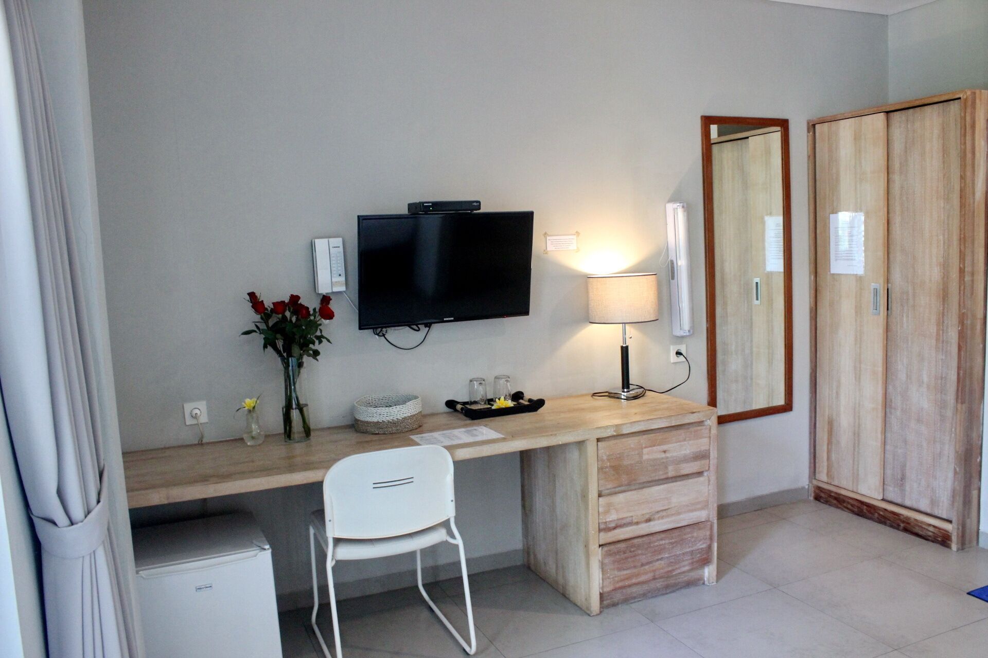 A hotel room with a wooden desk, a television, a lamp, a mirror, a small refrigerator, and a chair.