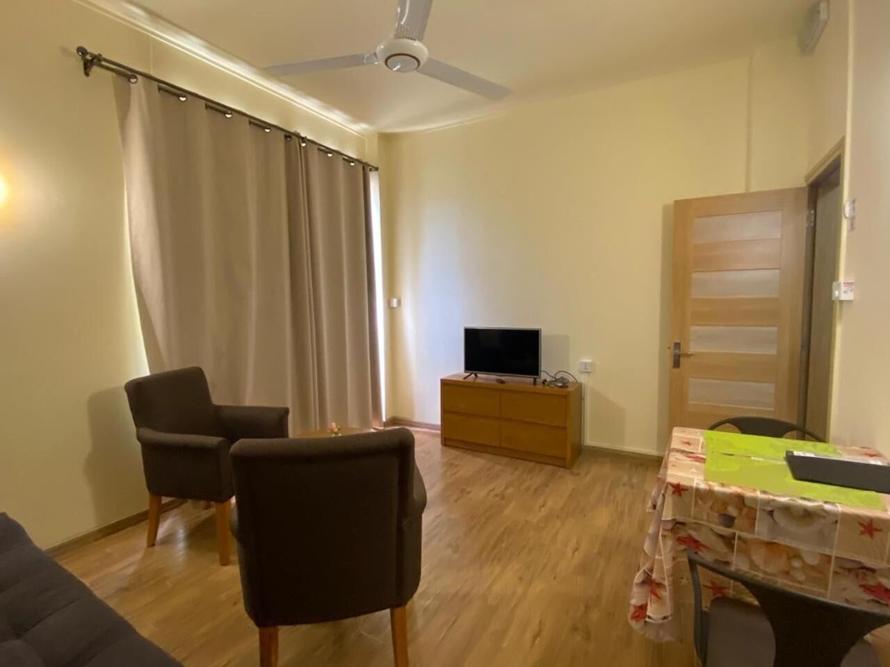 Un soggiorno con una ventola a soffitto, una televisione su un mobile in legno, due poltrone, un tavolo con tovaglia e una porta.