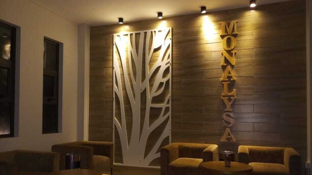 A reception area with a wooden wall, a decorative tree panel, and the name 'MONALYSA' in gold letters. There are two sofas and a round coffee table. - The Monalysa Suites & Residence (Grand-Baie)