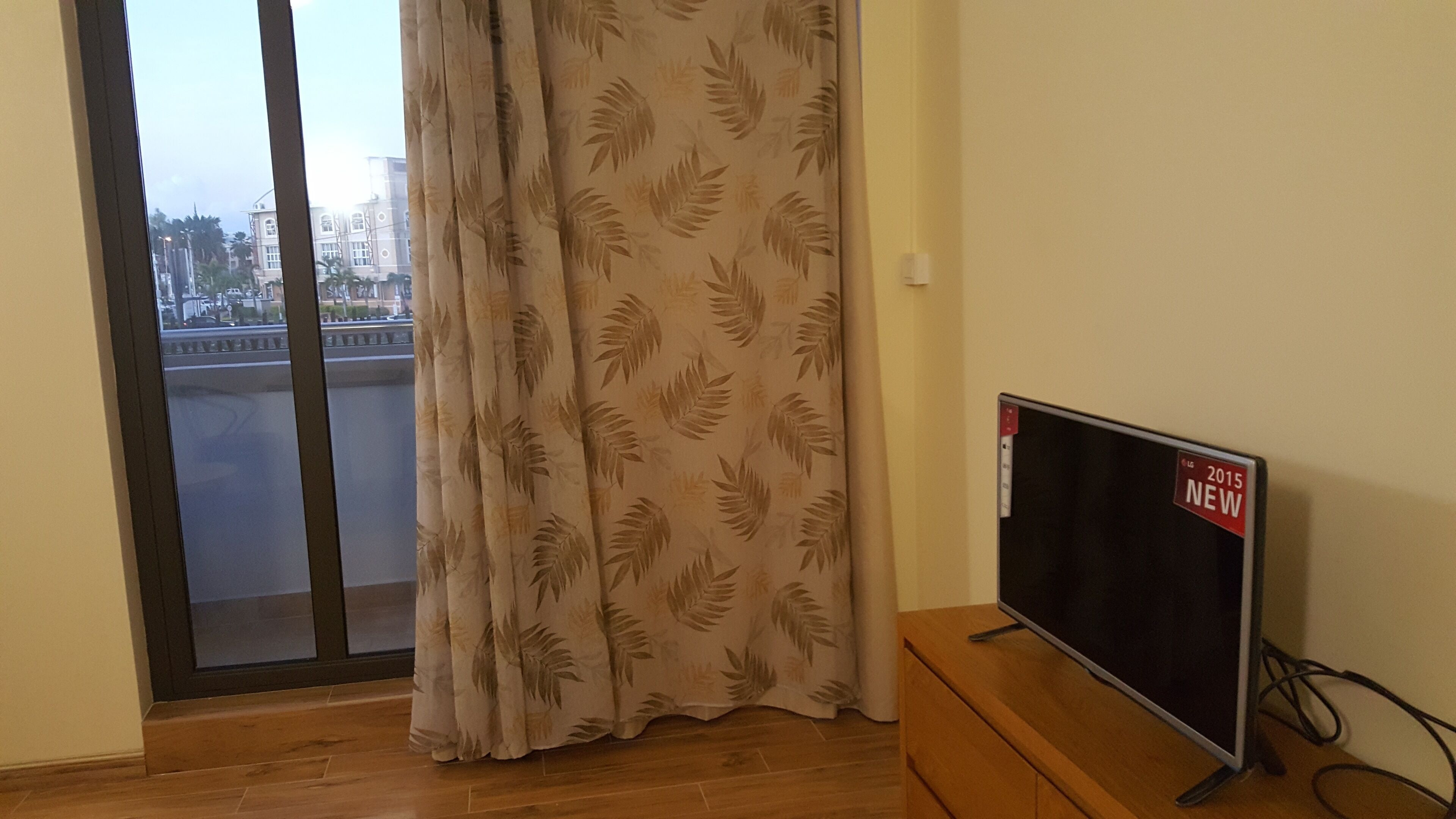 A room with a sliding glass door leading to a balcony, a flat-screen TV on a wooden stand, and a patterned curtain.