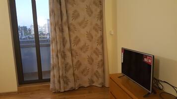 A room with a sliding glass door leading to a balcony, a flat-screen TV on a wooden stand, and a patterned curtain.