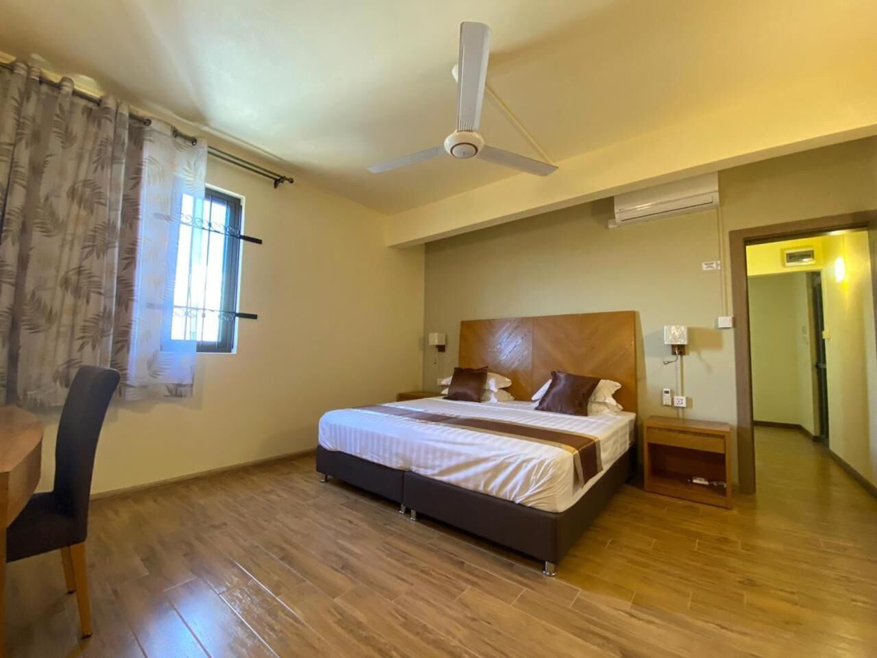Une chambre à coucher avec un lit, un ventilateur de plafond, une fenêtre avec des rideaux, un plancher en bois et une petite table avec une chaise.