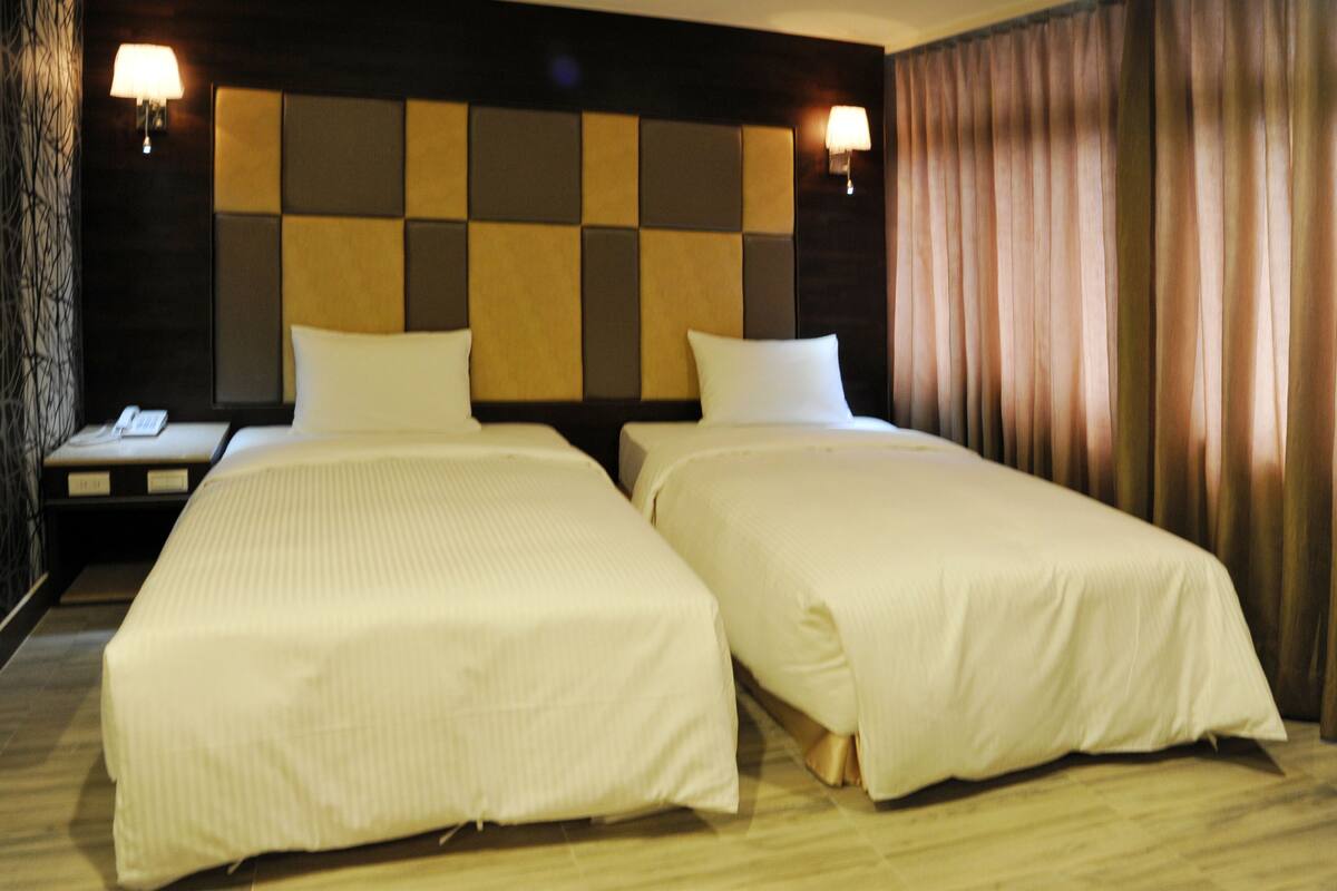 A hotel room with two single beds, a nightstand with a lamp, and a headboard with a square pattern.