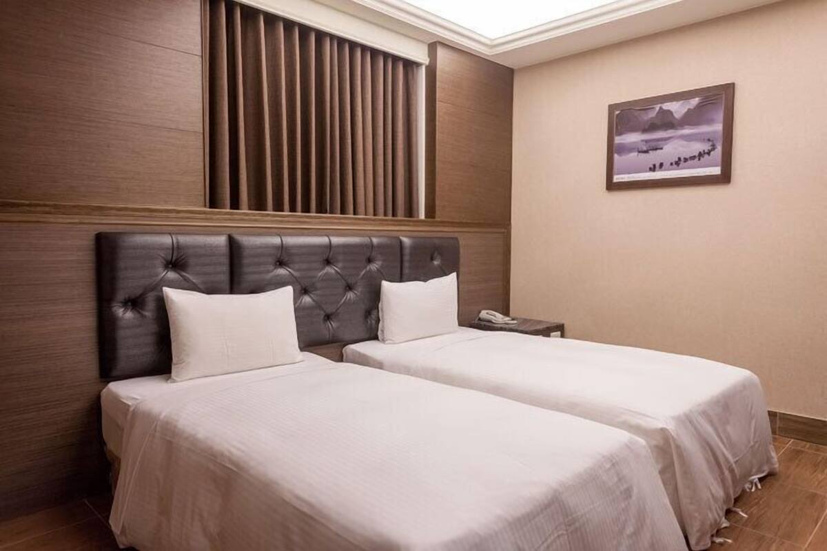A hotel room with two beds, a wooden headboard, a window with blinds, a framed picture on the wall, and a bedside table with a phone.