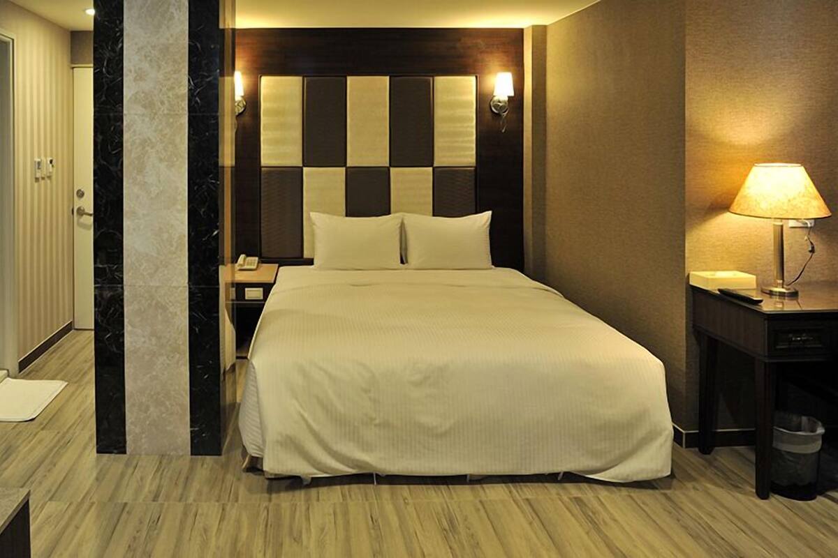 A hotel room with a large bed, a nightstand, a lamp, and a trash bin.