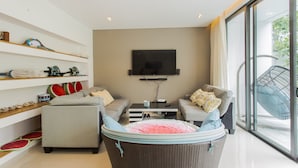 A modern living room with a sofa, armchair, and a wicker chair with a pink cushion. There are shelves with decorative items and a flat-screen TV mounted on the wall. - The Point Villa (Da Nang)
