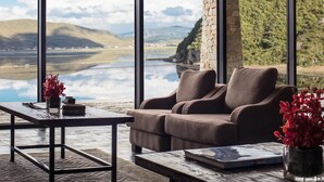 Een moderne woonkamer met een groot raam dat uitkijkt op een prachtig landschap, een comfortabele bank en een kleine tafel met een boek en bloemen.