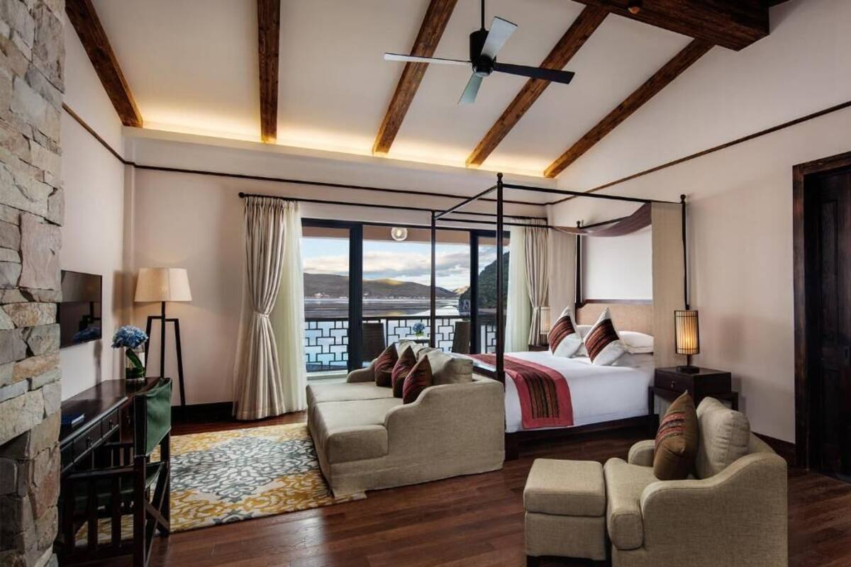 Une chambre à coucher avec un lit à baldaquin, un coin salon avec un canapé et un fauteuil, un bureau avec une lampe, et une vue sur l’extérieur grâce à de grandes fenêtres.