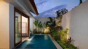 Une piscine dotée d’une terrasse en bois, entourée d’une végétation luxuriante et bordée d’un mur blanc.