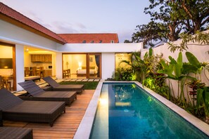 A modern outdoor pool area with lounge chairs, a wooden deck, and a kitchen visible through an open door.