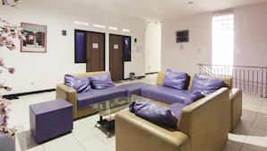 A modern living room with a sectional sofa, a glass coffee table, and a small side table. There are two doors with signs above them, and a window with a view of the outside. - Ungu Residence Syariah (Bandung)