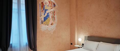 A bedroom with a large bed, wooden flooring, a nightstand, and a wall with a decorative painting.