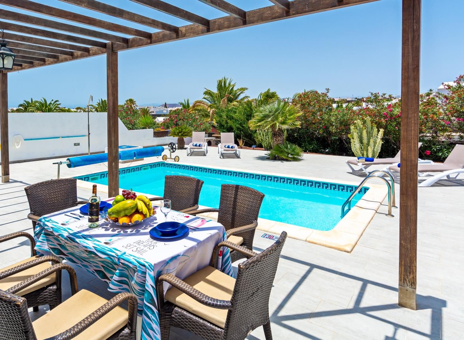 Zona junto a la piscina con mesa lista para comer, sillas de descanso y una estructura de pérgola.