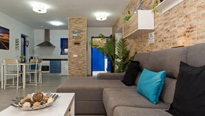 Een moderne woonkamer met een grijze bank, een witte salontafel en een blauw kussen. Achtergrond is een keuken met blauwe kastjes en een bakstenen muur.