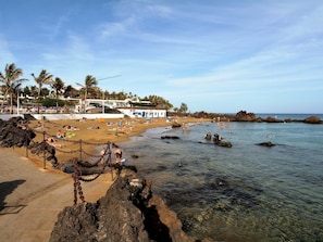 A beach with clear water, people swimming, and a chain-link fence separating the sand from the water. - Playa Grande Puerto de Carmen Share Pool By PVL (Puerto del Carmen)