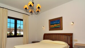 A bedroom with a wooden bed, a window with curtains, a chandelier, a bedside table, a nightstand, a wall-mounted lamp, and a framed picture. - Villa Timanfaya Private Pool By PVL (Yaiza)