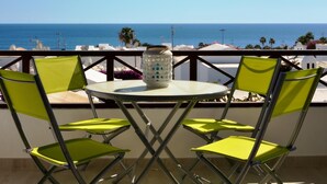A balcony with a round table and four green chairs, overlooking a coastal view with buildings and palm trees.
