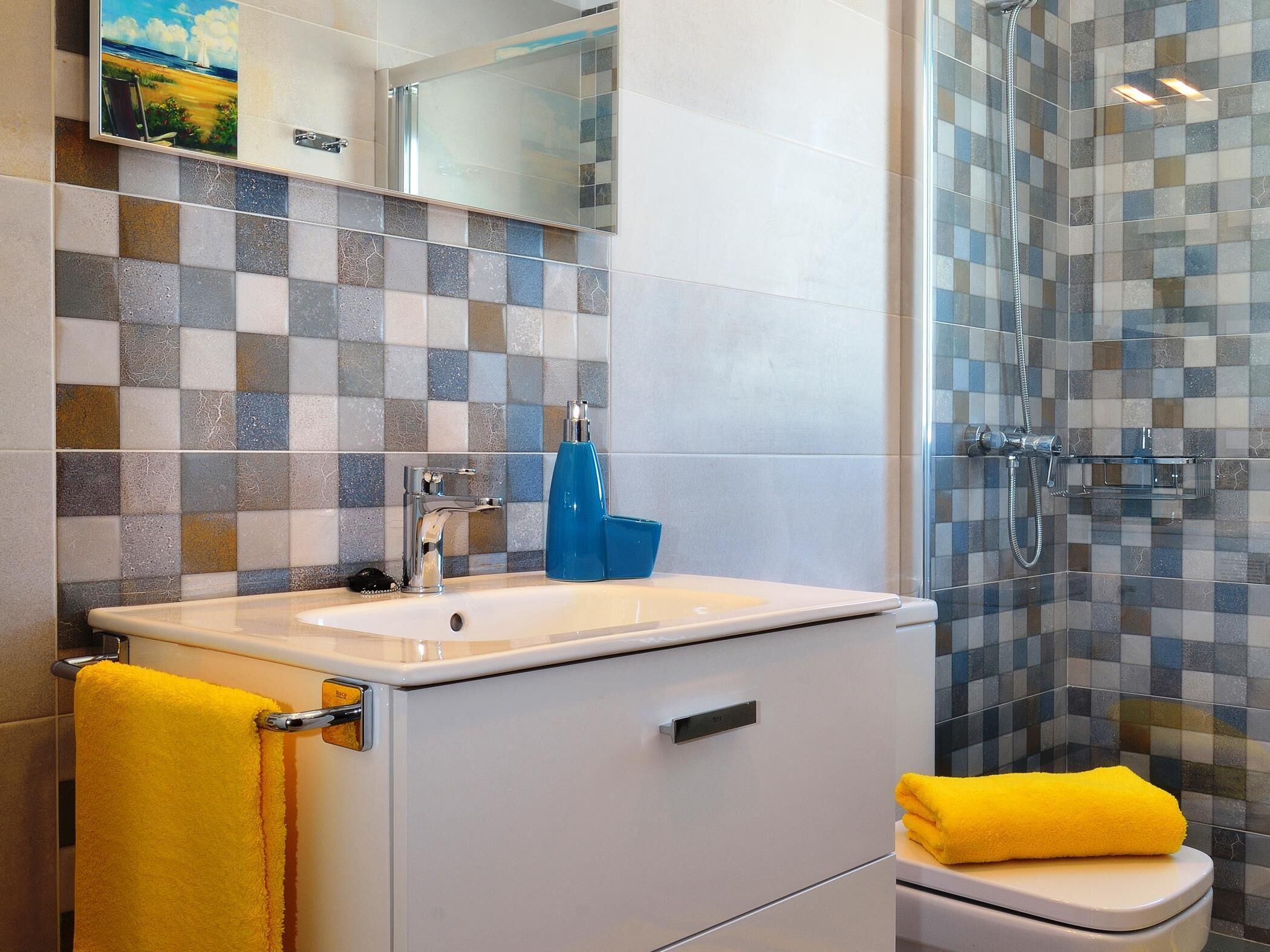A bathroom with a tiled shower wall, a sink with a blue soap dispenser, and a yellow towel on the floor.