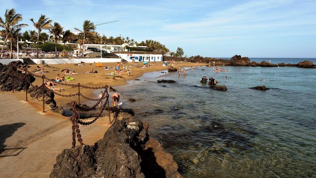 A beach with clear water, people swimming, and a chain-link fence separating the sand from the water.