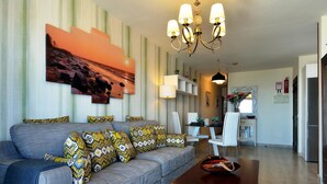 A modern living room with a grey sofa, patterned cushions, a wooden coffee table, and a large painting of a sunset.