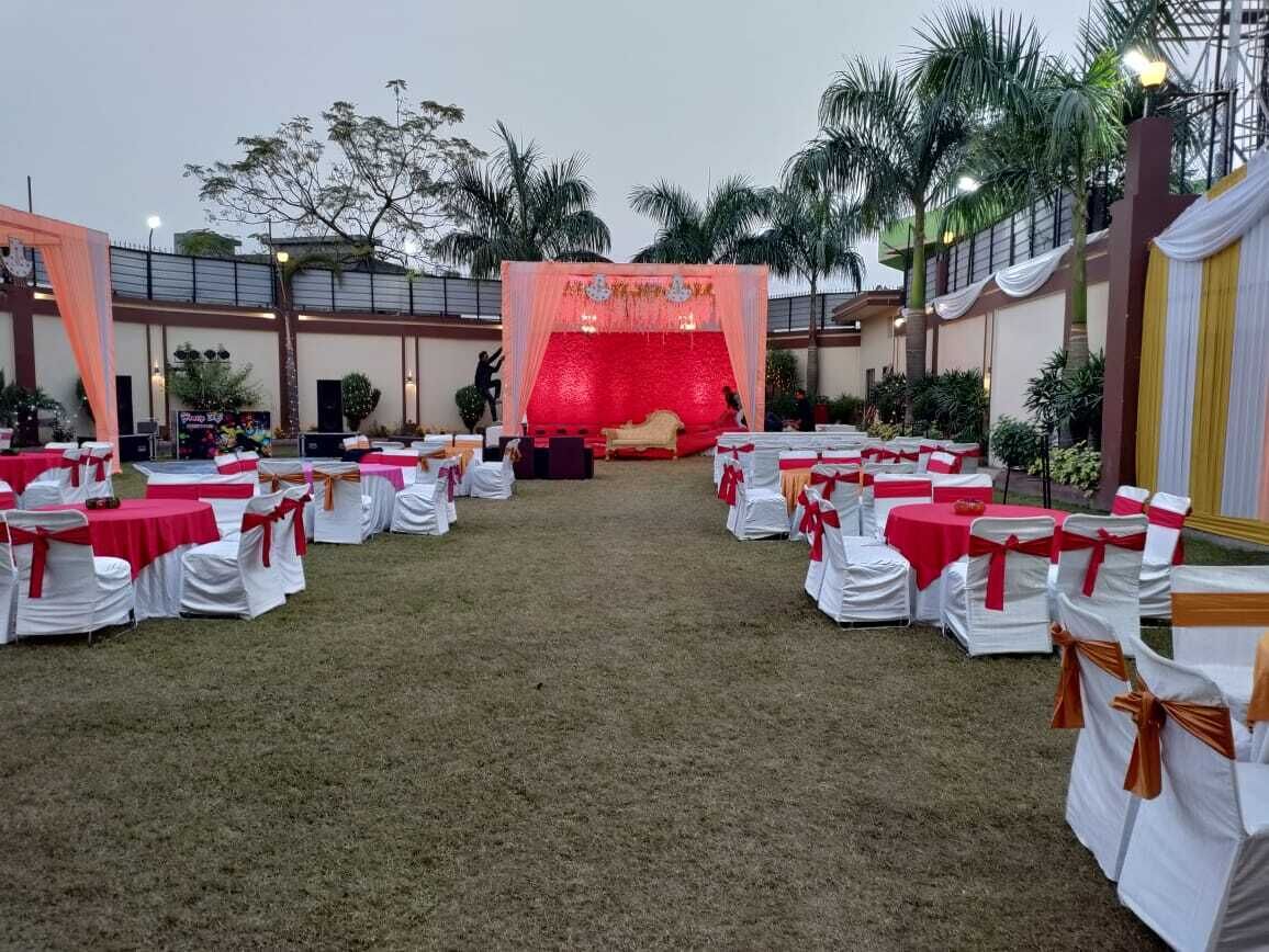 Un espacio para eventos al aire libre con un escenario, filas de mesas blancas con lazos rojos y anaranjados, y palmeras al fondo.