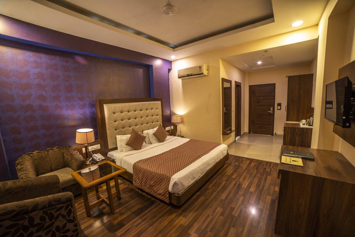 A hotel room with a large bed, a sofa, a coffee table, an air conditioning unit, a TV, and a mirror.
