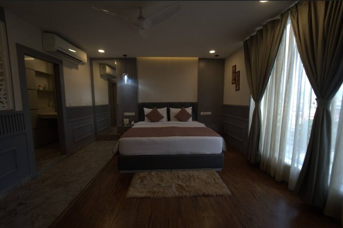 A bedroom with a large bed, a wooden floor, an air conditioning unit, and a window with curtains.