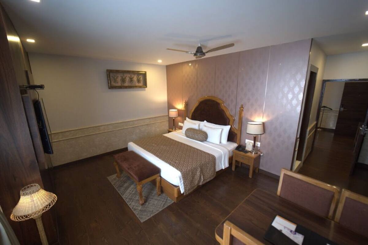 A hotel room with a large bed, a wooden bench, a desk with chairs, a ceiling fan, and a wall-mounted TV.